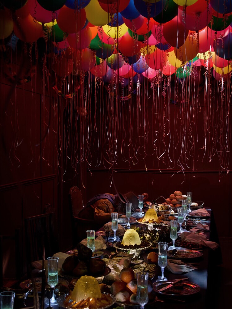 A long dining table filled with food and glasses, a collection of colourful balloons with long strings attached hang above, a person is slumped over the table on a chair in the far left corner  
